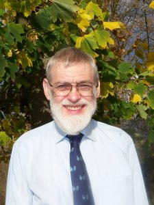 Peter Loader head shot. Peter wears a shirt and tie and stands infront of a background with green and yellow leaves. 