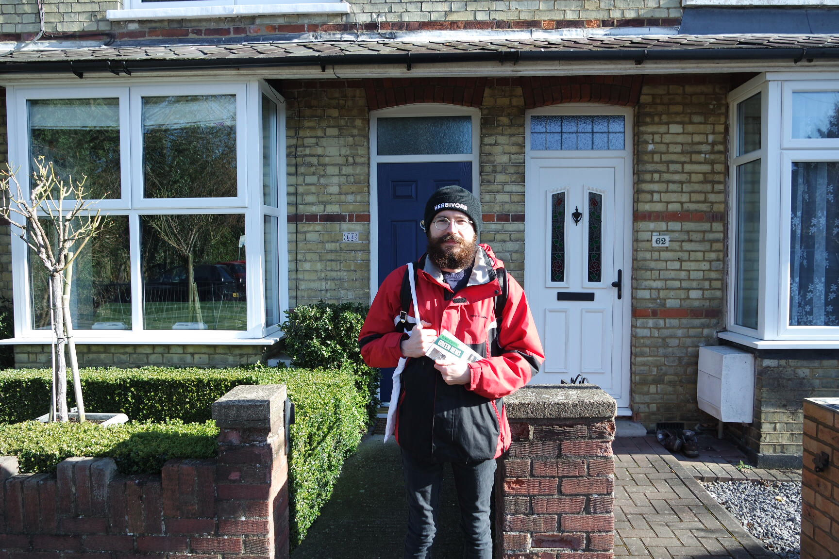 Tom pashby stands outside terraced houses holding leaflets and wearing a hat and coat. The sun is shining from the right of the frame, creating highlight and shadow.