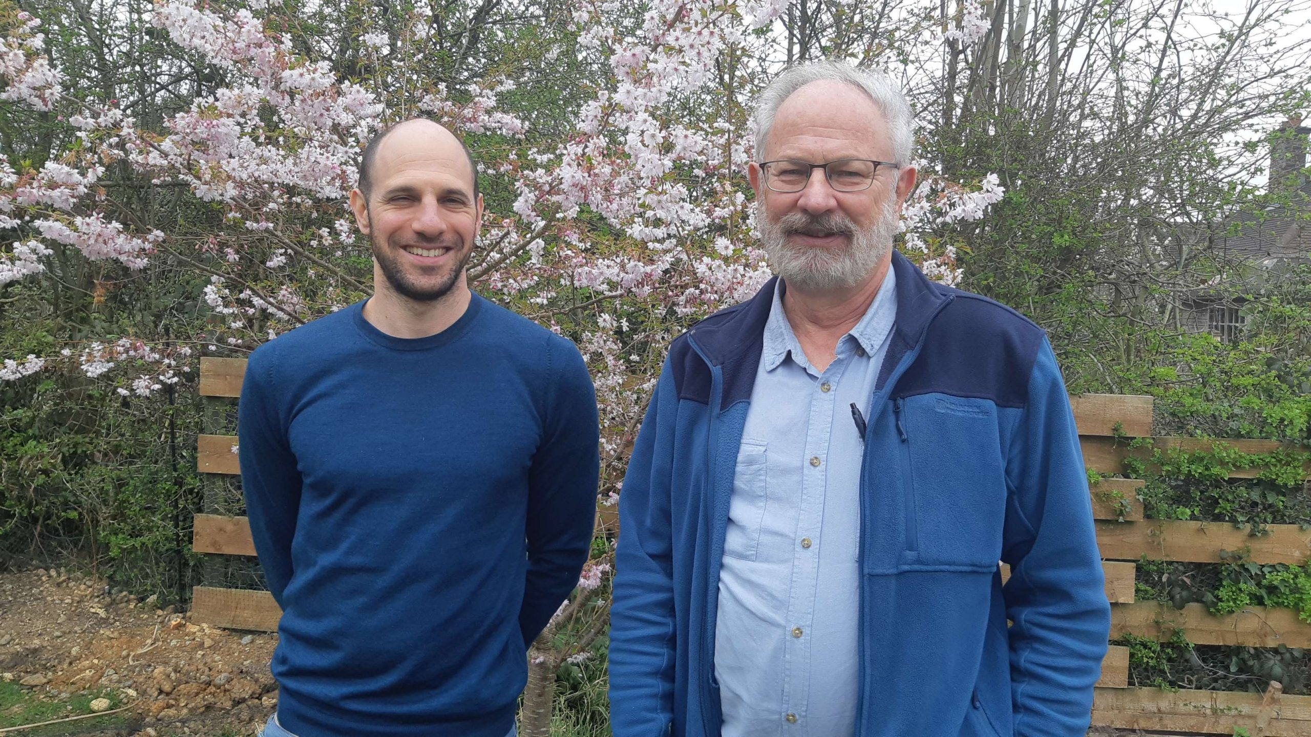 Alex Micheals stands next to Chris Mitchell. Both are smiling and standing in front of a tree in blossom