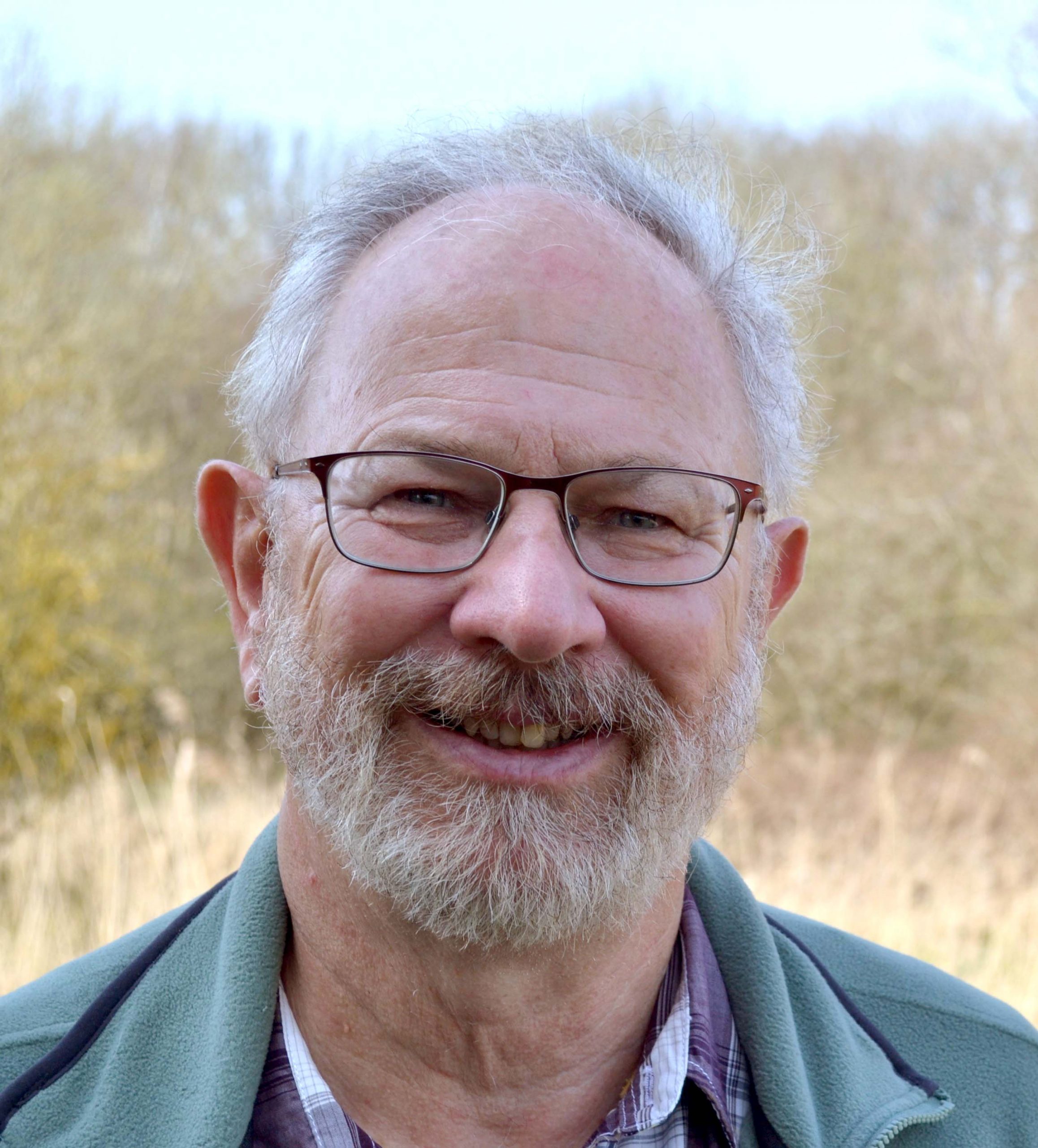 Head shot of Chris Mitchell. He is standing infront of a natural landscape, with short grey hair and a beard. He is wearing a blue coat.