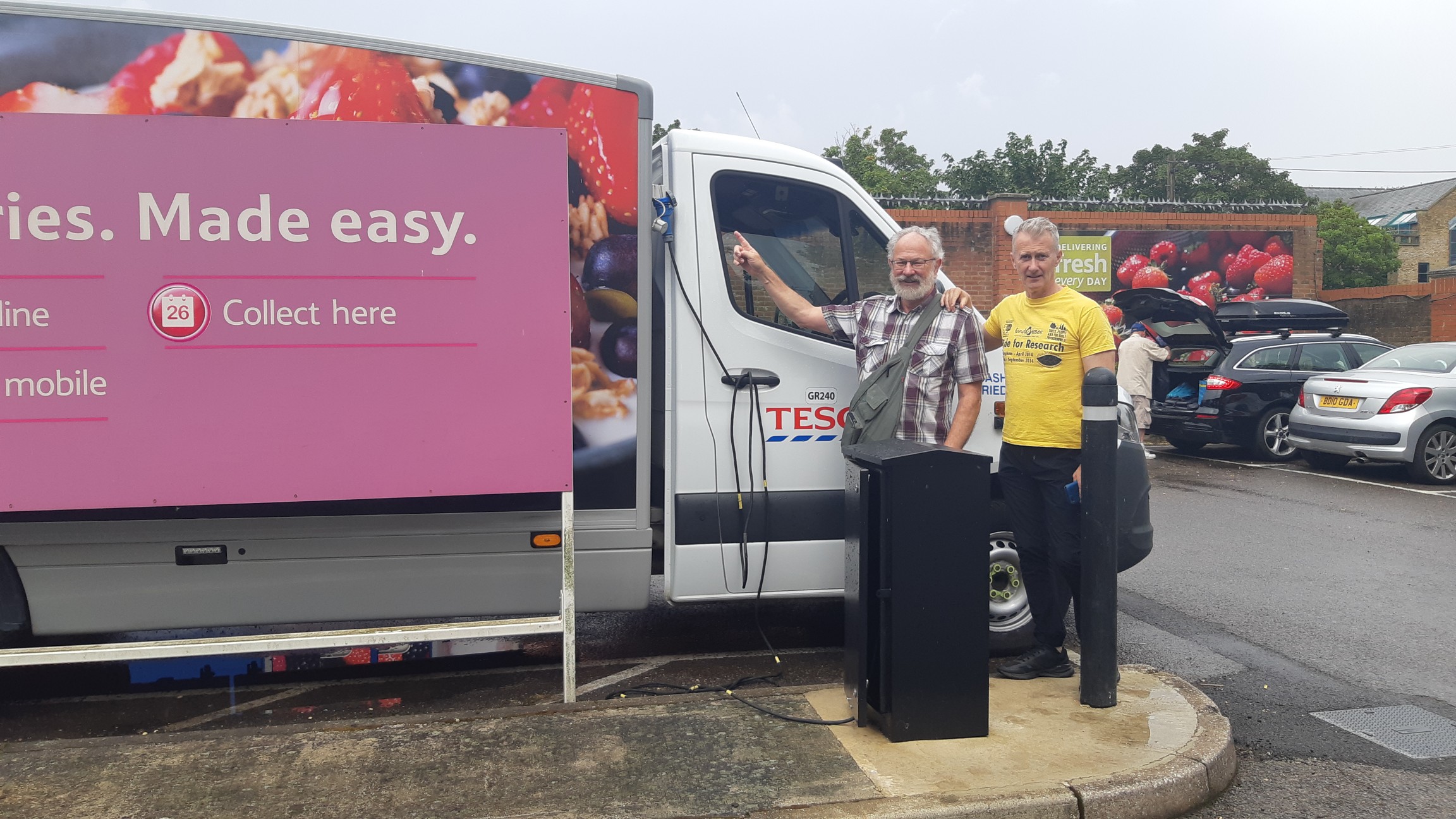 Two men stand in front of a Tesco delivery van. One is pointing to a new charging cable connected to the van